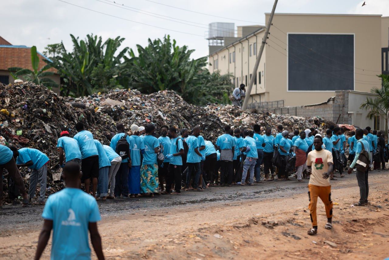 A long line of people in light blue shirts along the face of the waste pile, some bending to the waste, buildings and banana trees behind