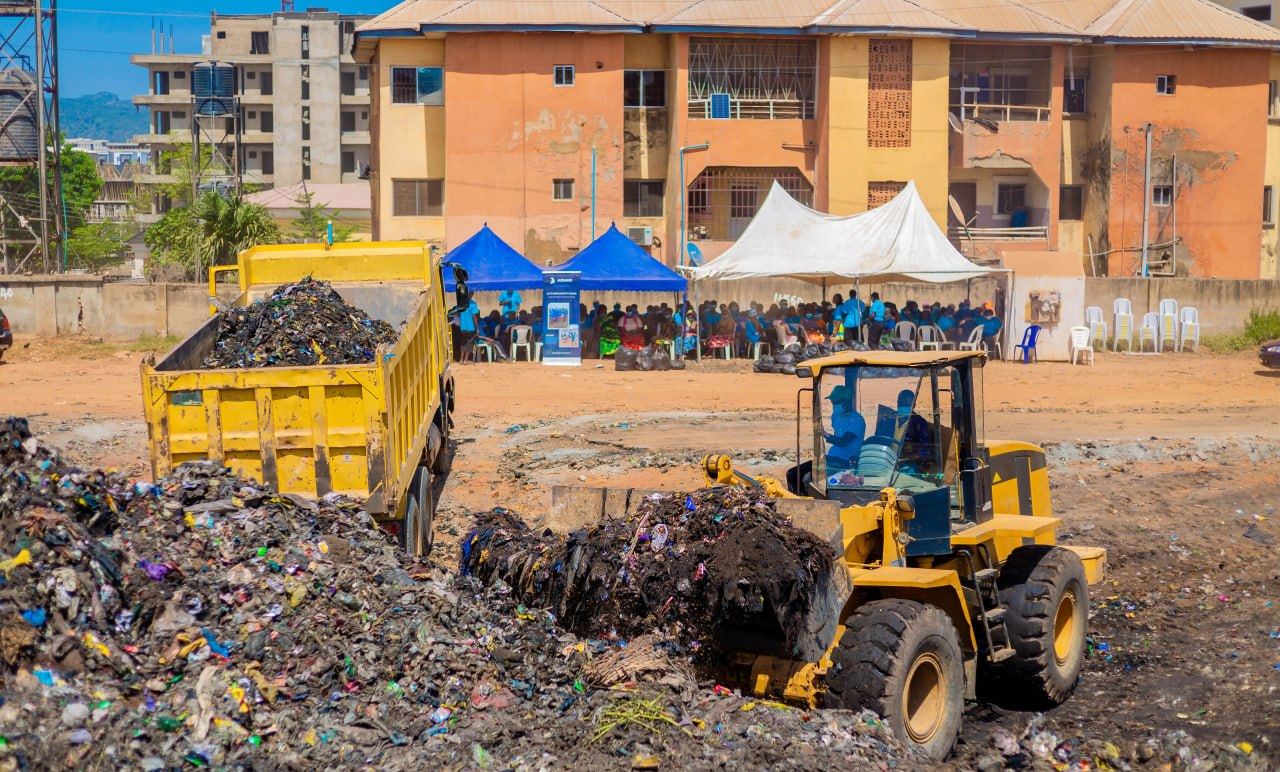 A yellow wheel loader filling a yellow tipper truck with waste; residents seated under blue and white tents behind